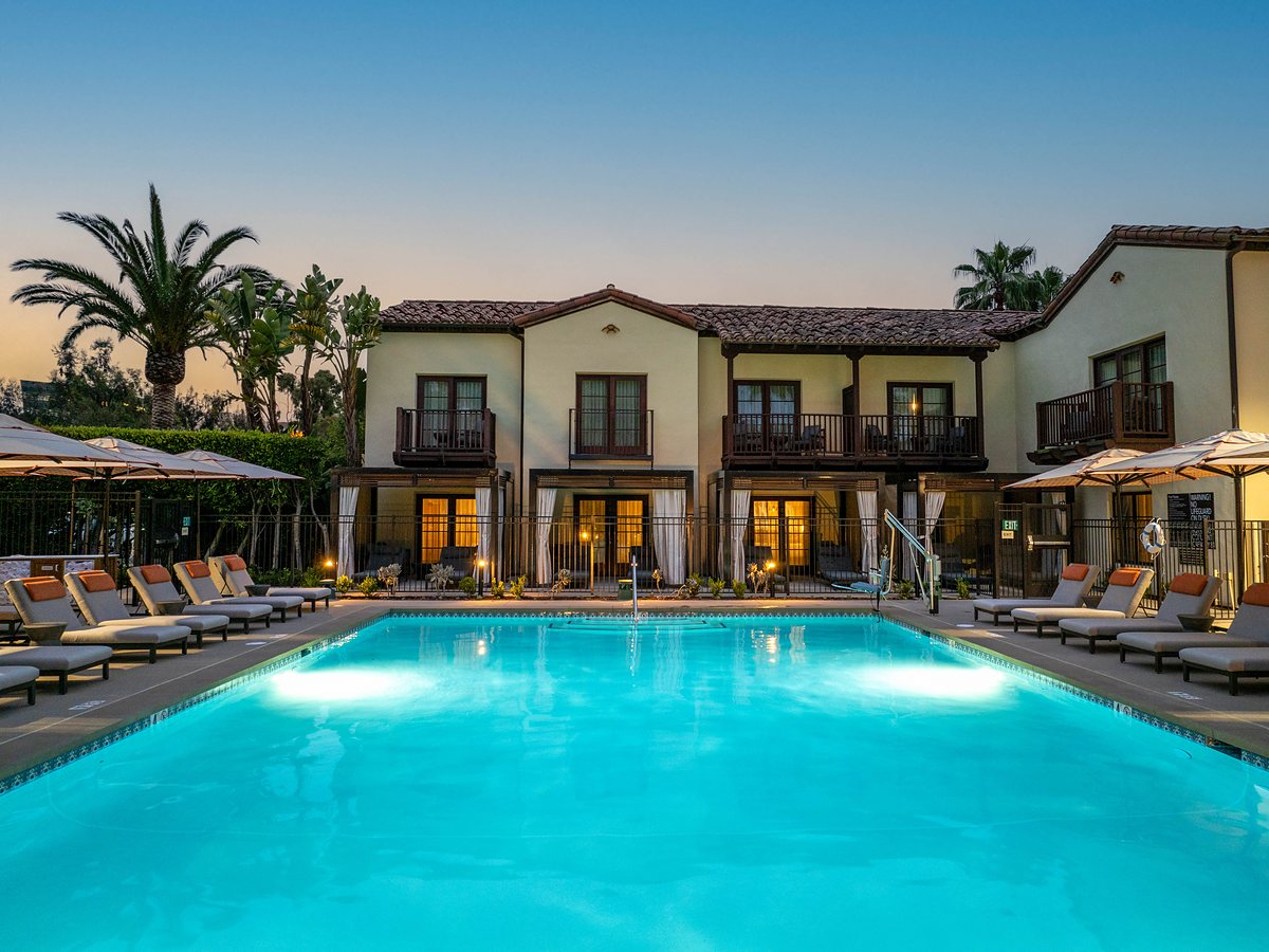 Illuminated swimming pool and lounge chairs at dusk surrounded by Spanish hacienda-style guest rooms with palm trees at Estancia La Jolla resort in La Jolla, California