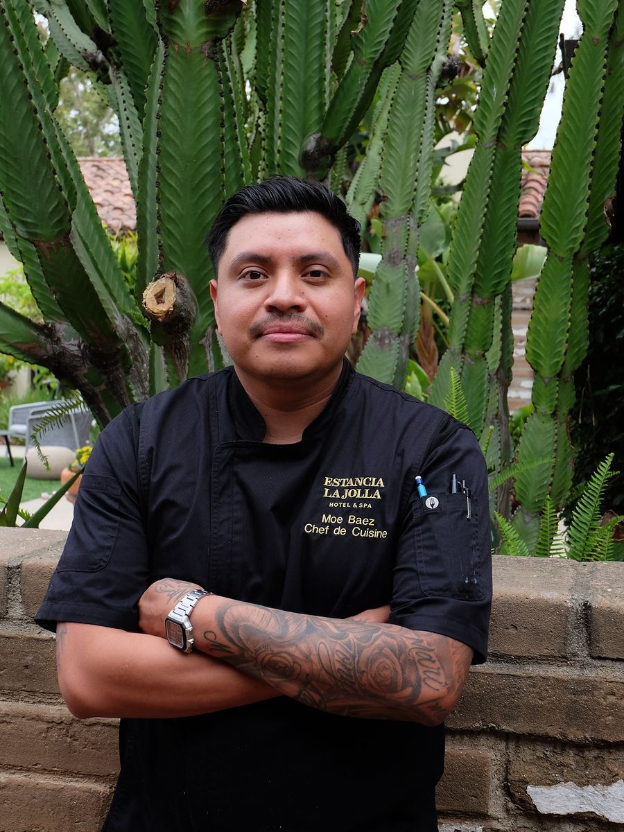 Chef de Cuisine Moe Baez smiling in his chef coat against a lush cactus backdrop at Estancia La Jolla Hotel & Spa in La Jolla, California.